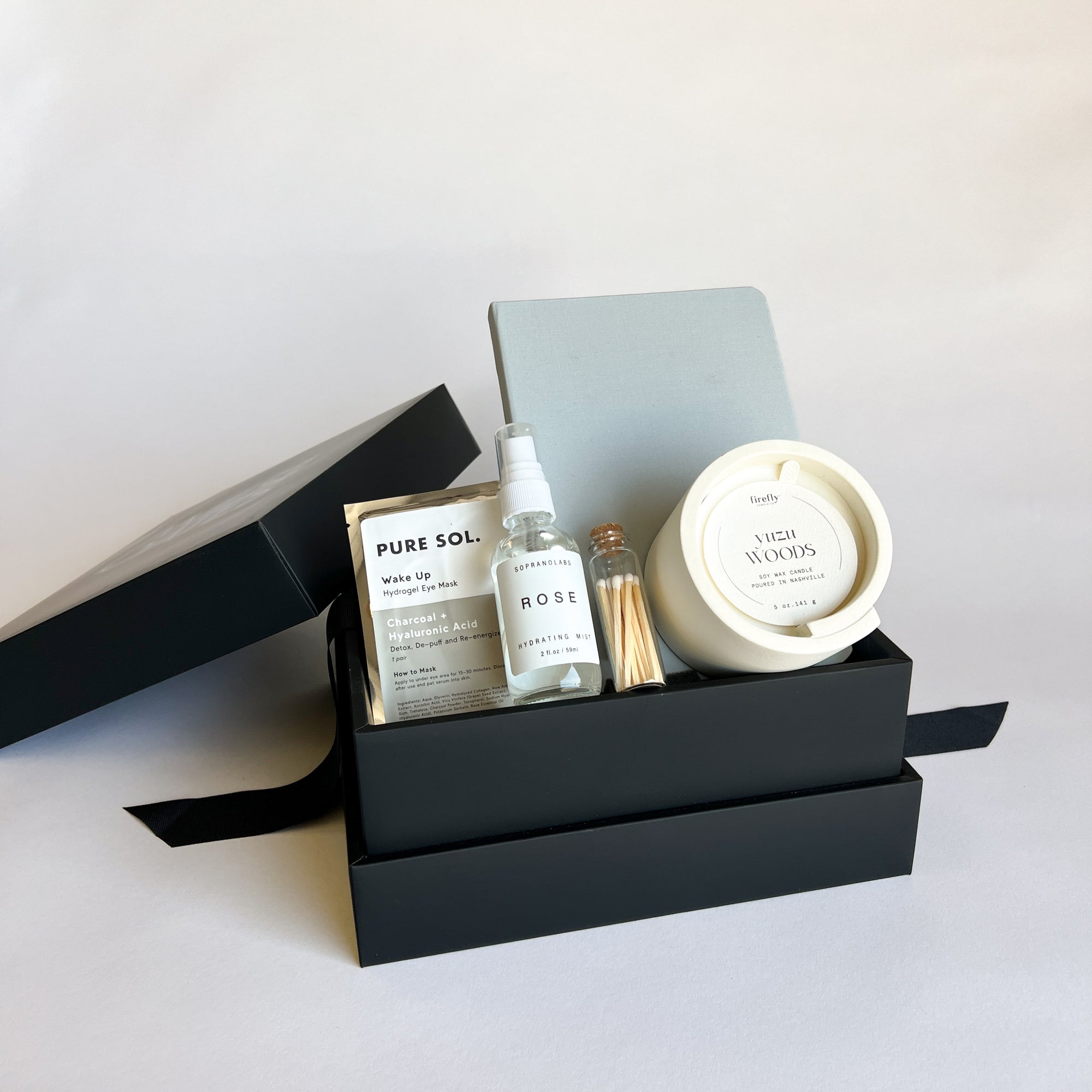 A gift box open to display its contents, which include a rose hydrating mist, a gray hardcover notebook, a soy wax candle in a concrete vessel, matches in an apothecary jar, and under eye hydrogel patches.