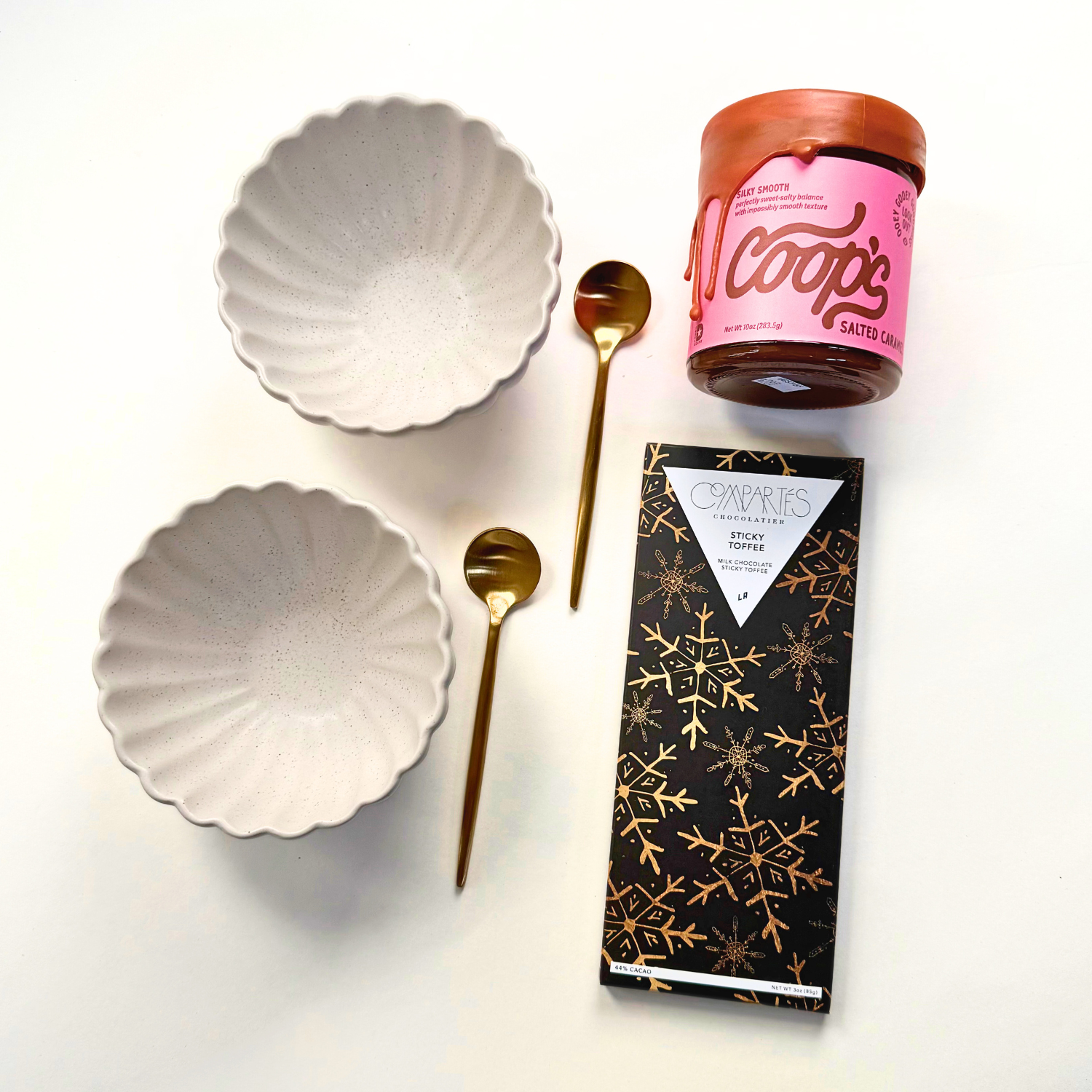 Two bowls, two gold spoons, coop's salted caramel sauce and compartes chocolate bar on white background