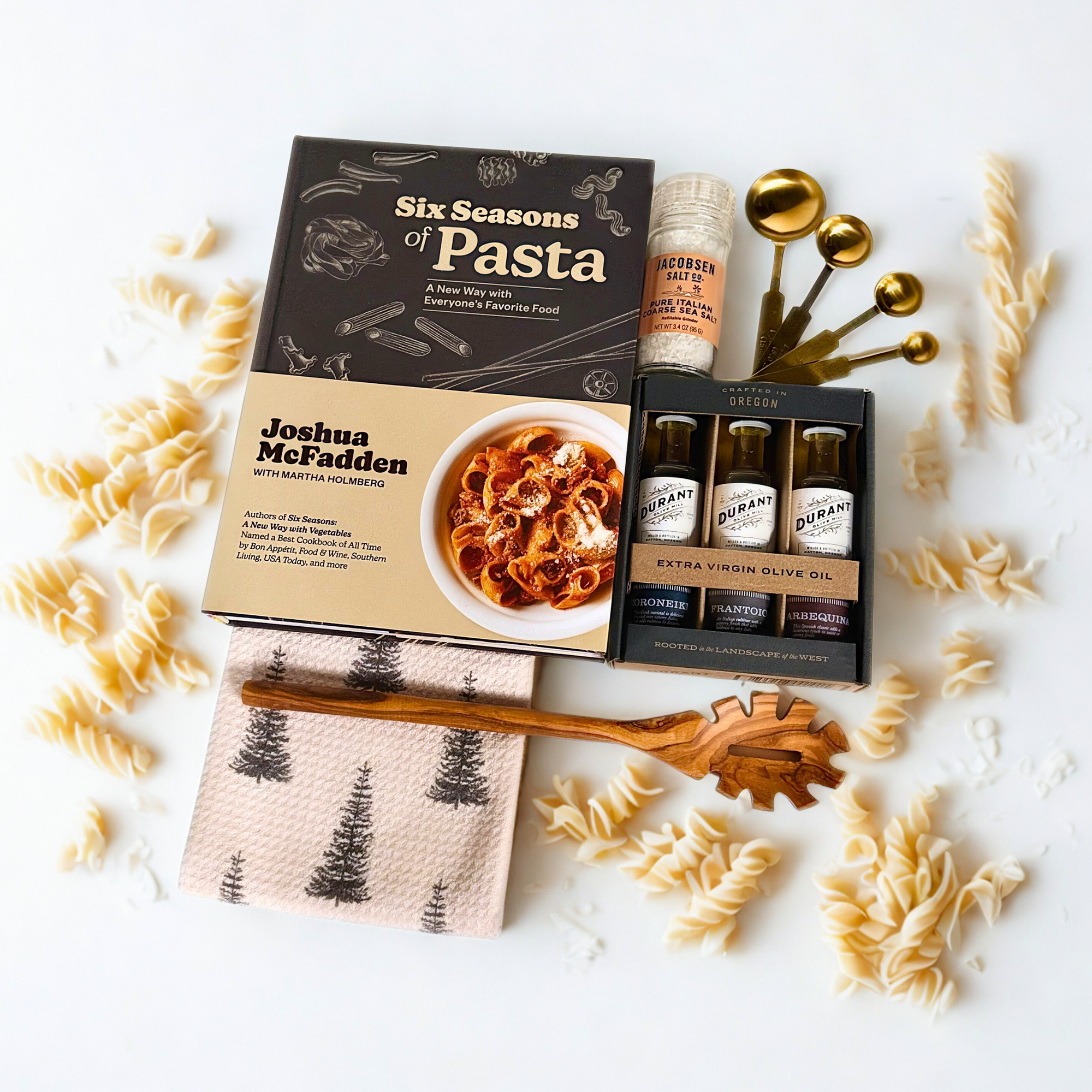 Pasta kit with 'Six Seasons of Pasta' book, Duran olive oil bottles,  jacobsen italian salt, tea towel, olive wood pasta spoon, gold measuring spoons on a white surface.