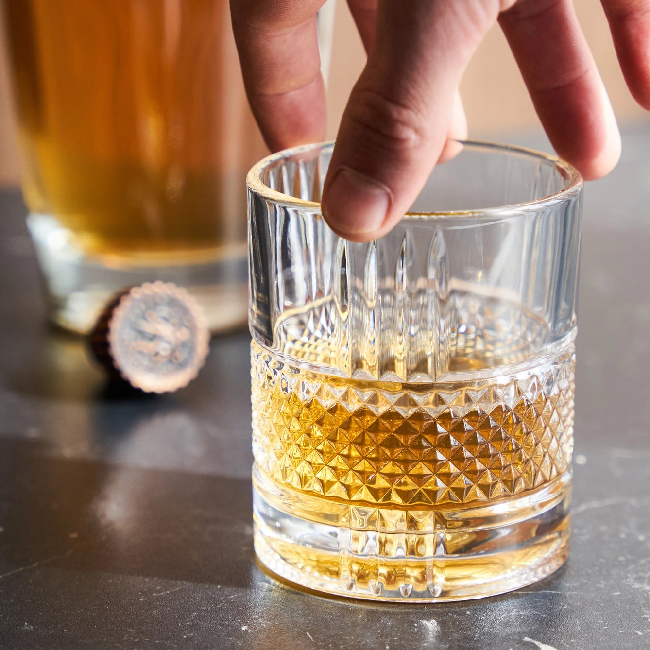 Glass of whiskey with a hand reaching for it on a dark surface