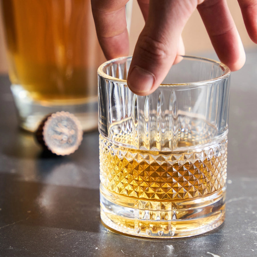 Collection of items including a bottle of Jack Rudy Cocktail Co., a revolving glass, and a box of Daneson toothpicks, artisan playing cards, and dark chocolate on a white surface.
