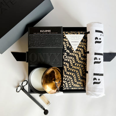 A gift box containing various items including a chocolate bar, a candle, a wick trimmer, and a cloth towel, all displayed in front of a black background.